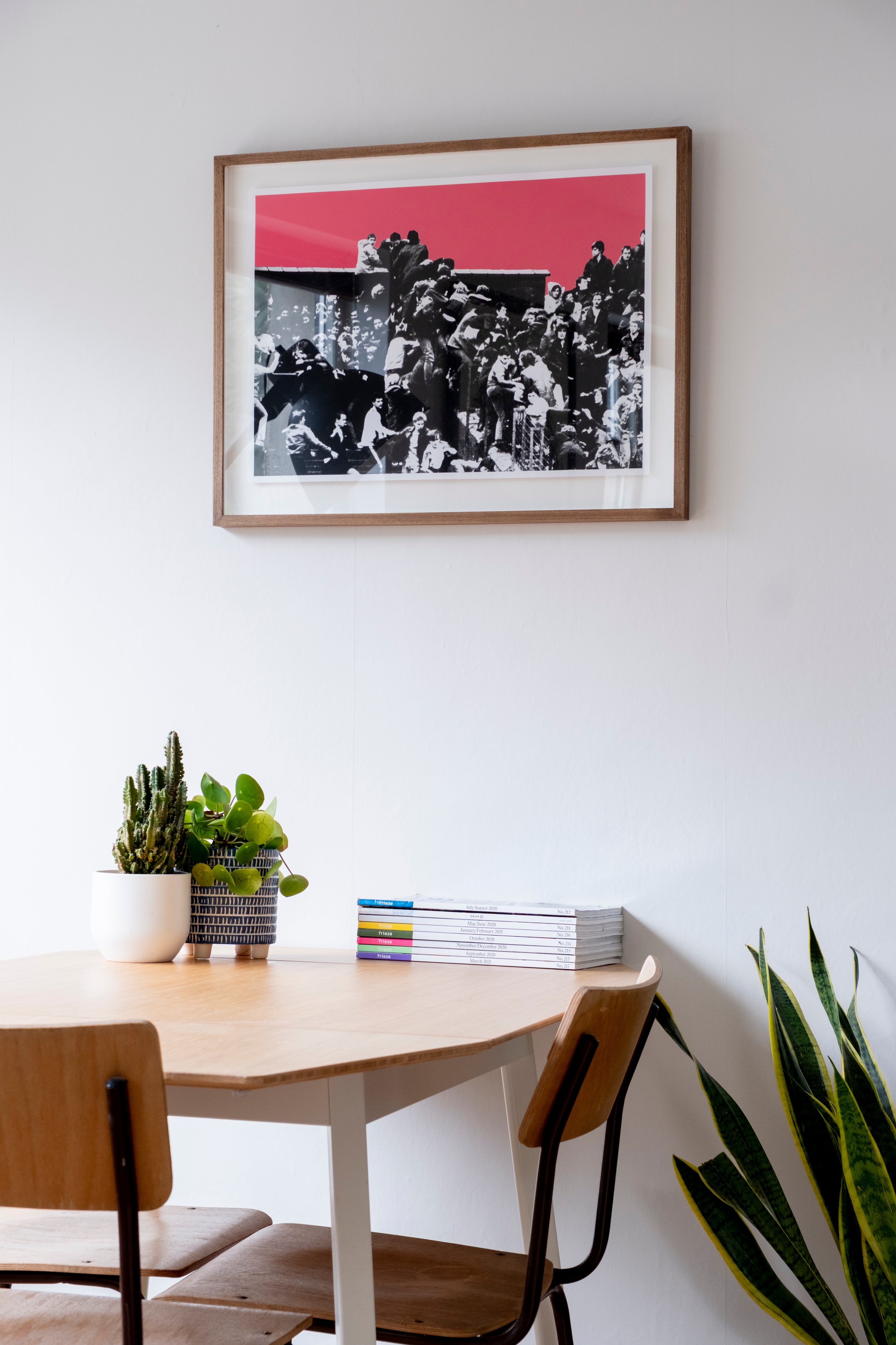 Framed artwork above a dining table with chairs and plants in a room.
