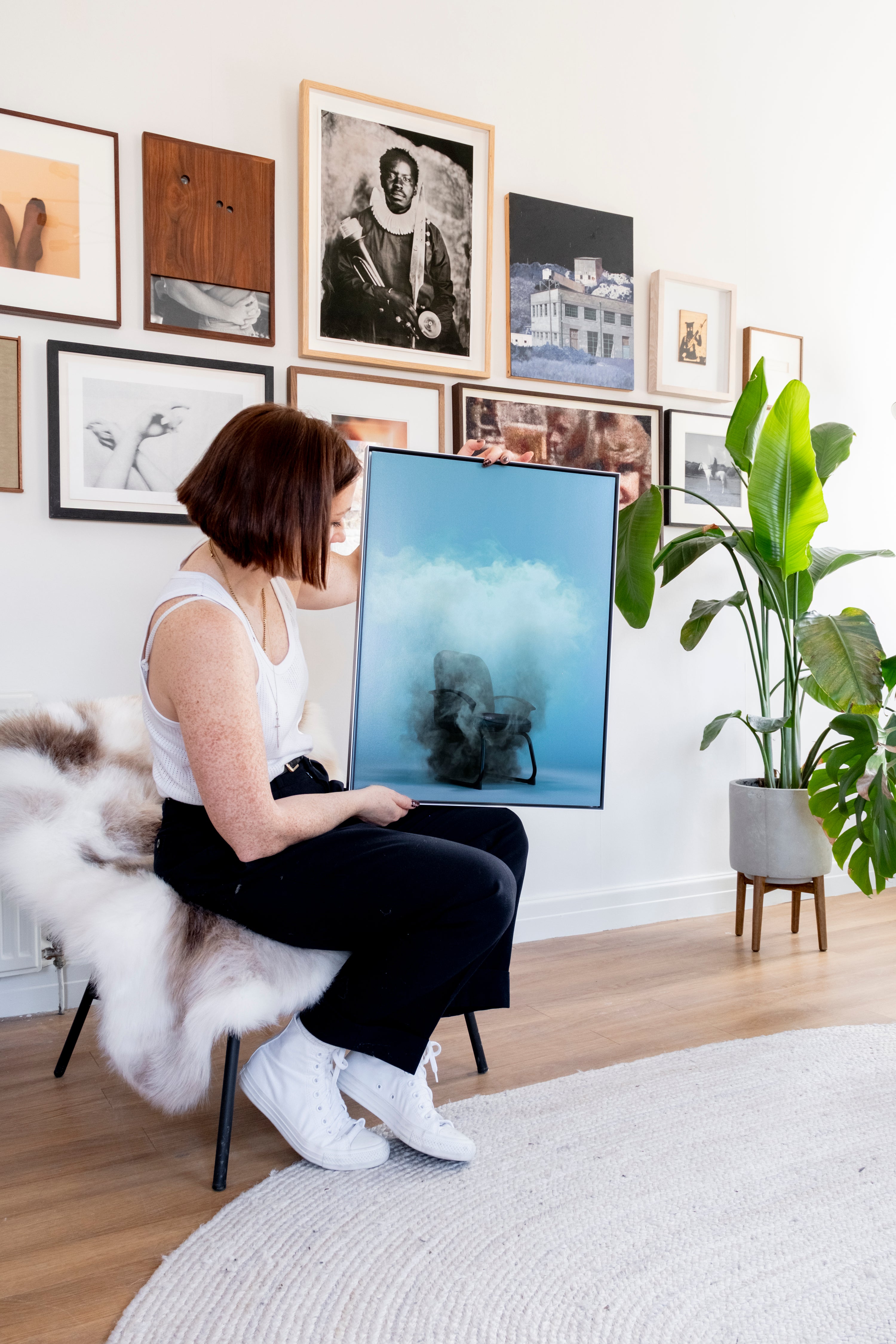 Woman sitting on a stool in a room with framed pictures on the wall, holding a large screen displaying an image.