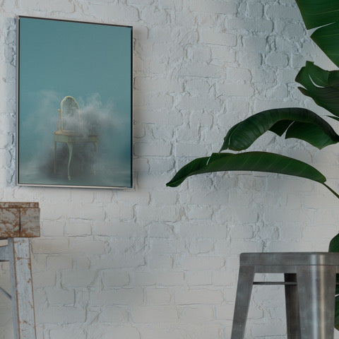 Framed artwork on a white brick wall with a plant and metal stool in the foreground.